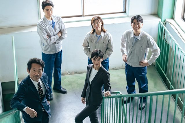 作業着やスーツ姿の5人が、階段の踊り場でこちらを見上げている様子